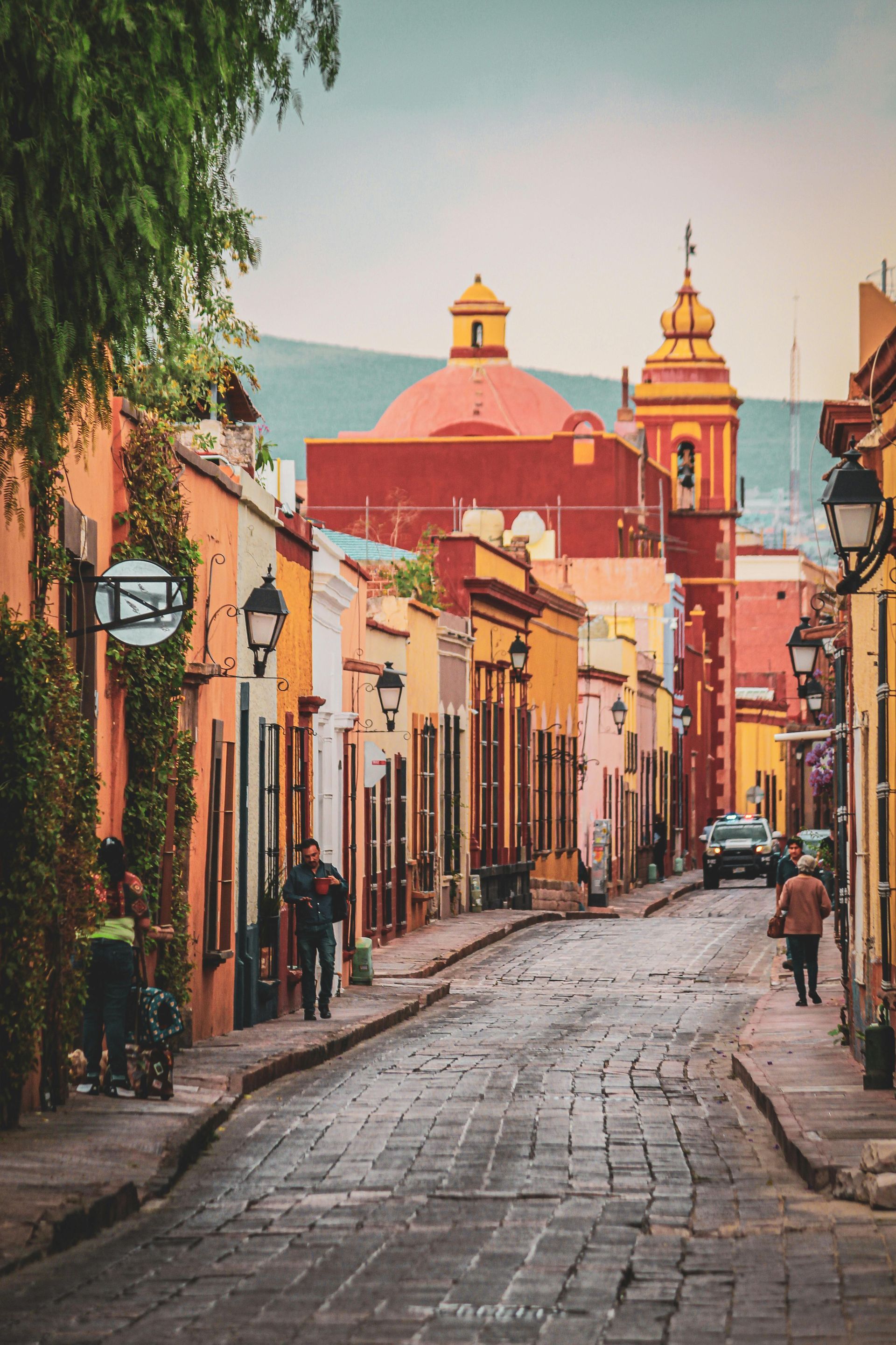 Cobblestone street in San Miguel de Allende, Mexico, lined with colorful buildings; people walk towards a church.