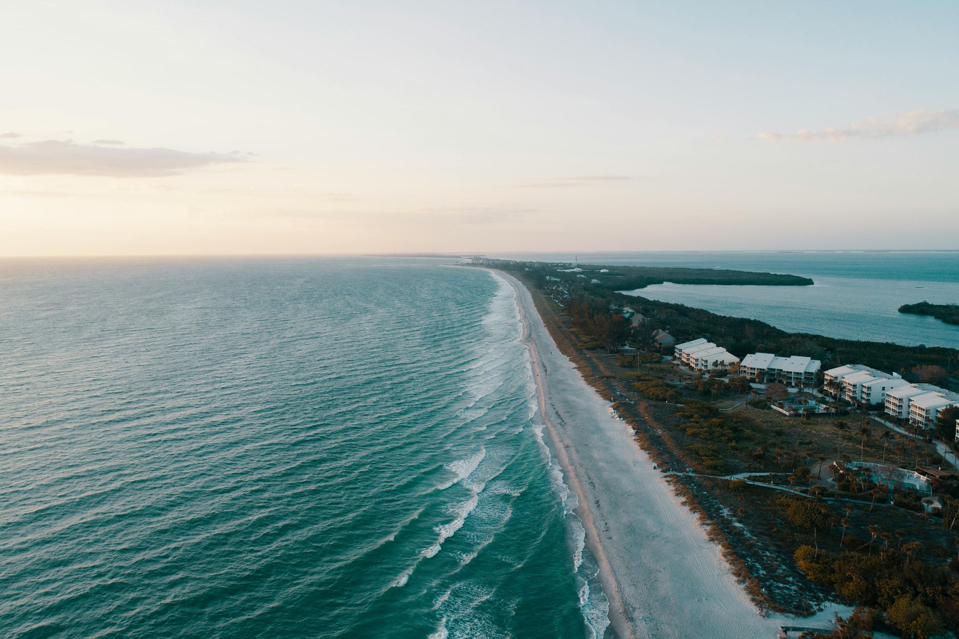 Aerial view of a long beach, turquoise water, and a strip of land with buildings along the shore under a soft sky.