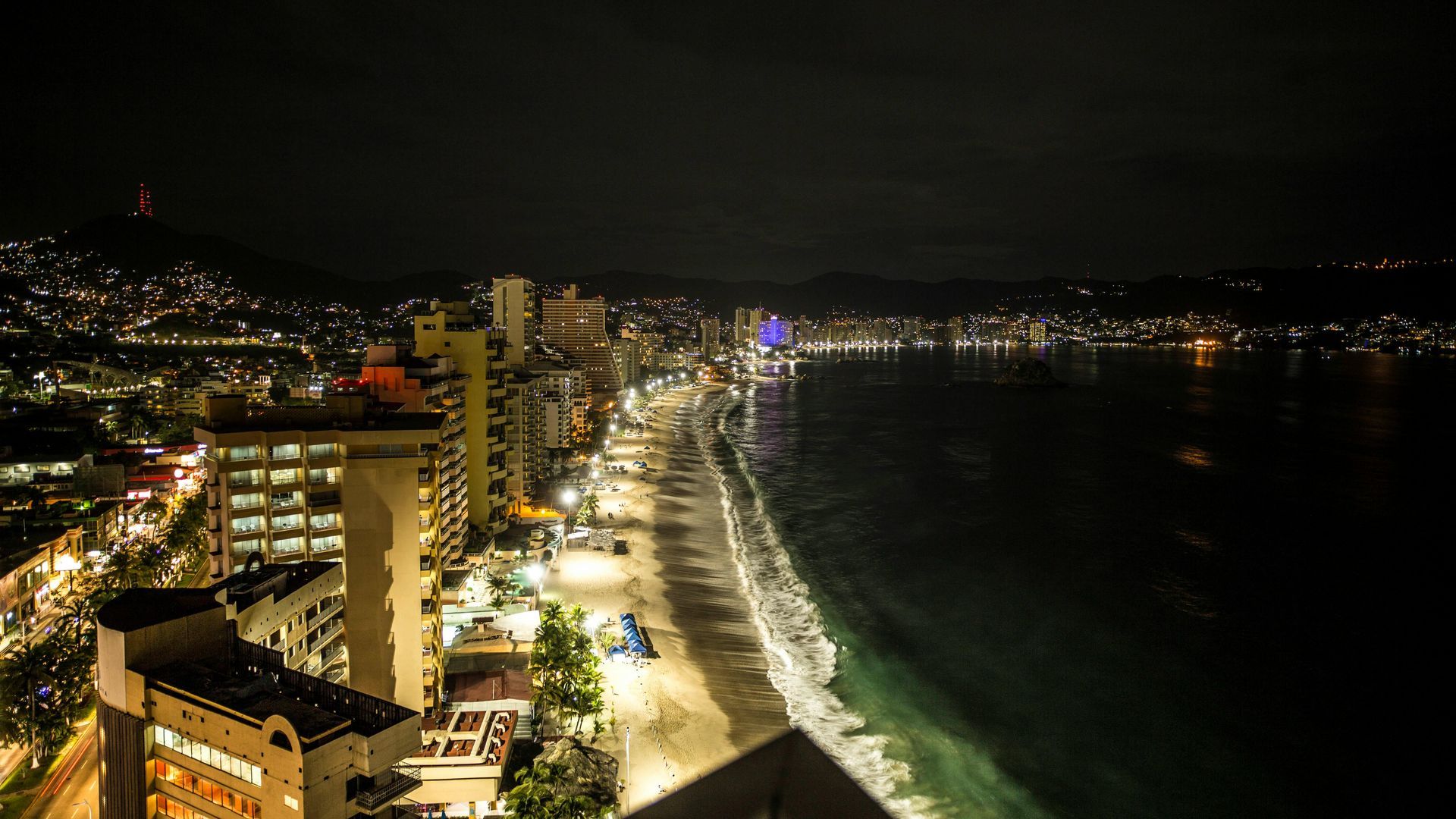 Night view of a beachside city with lights lining the shore and along buildings.