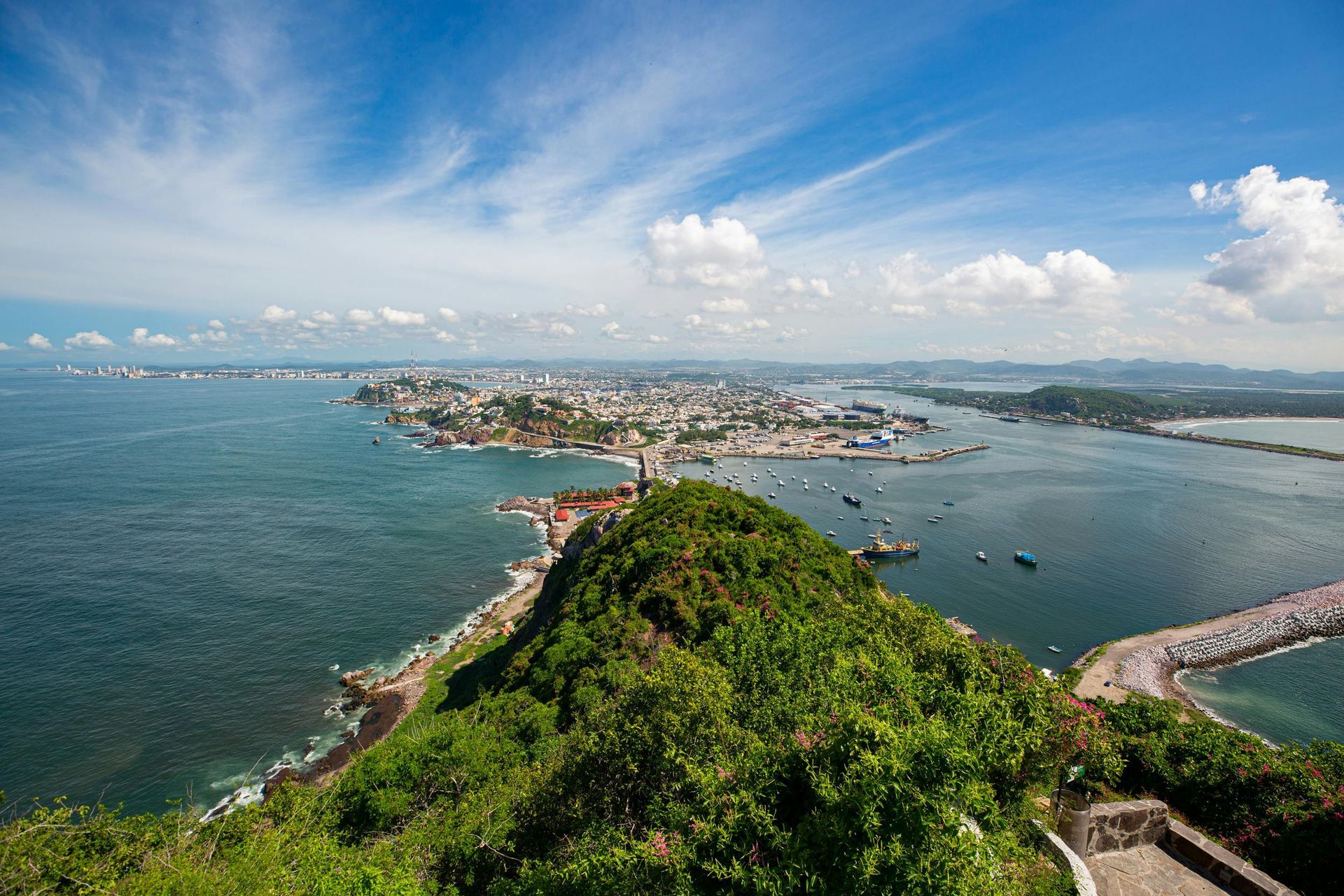 Scenic view of a coastal city on a peninsula with blue water, a cloudy sky, and lush green vegetation.