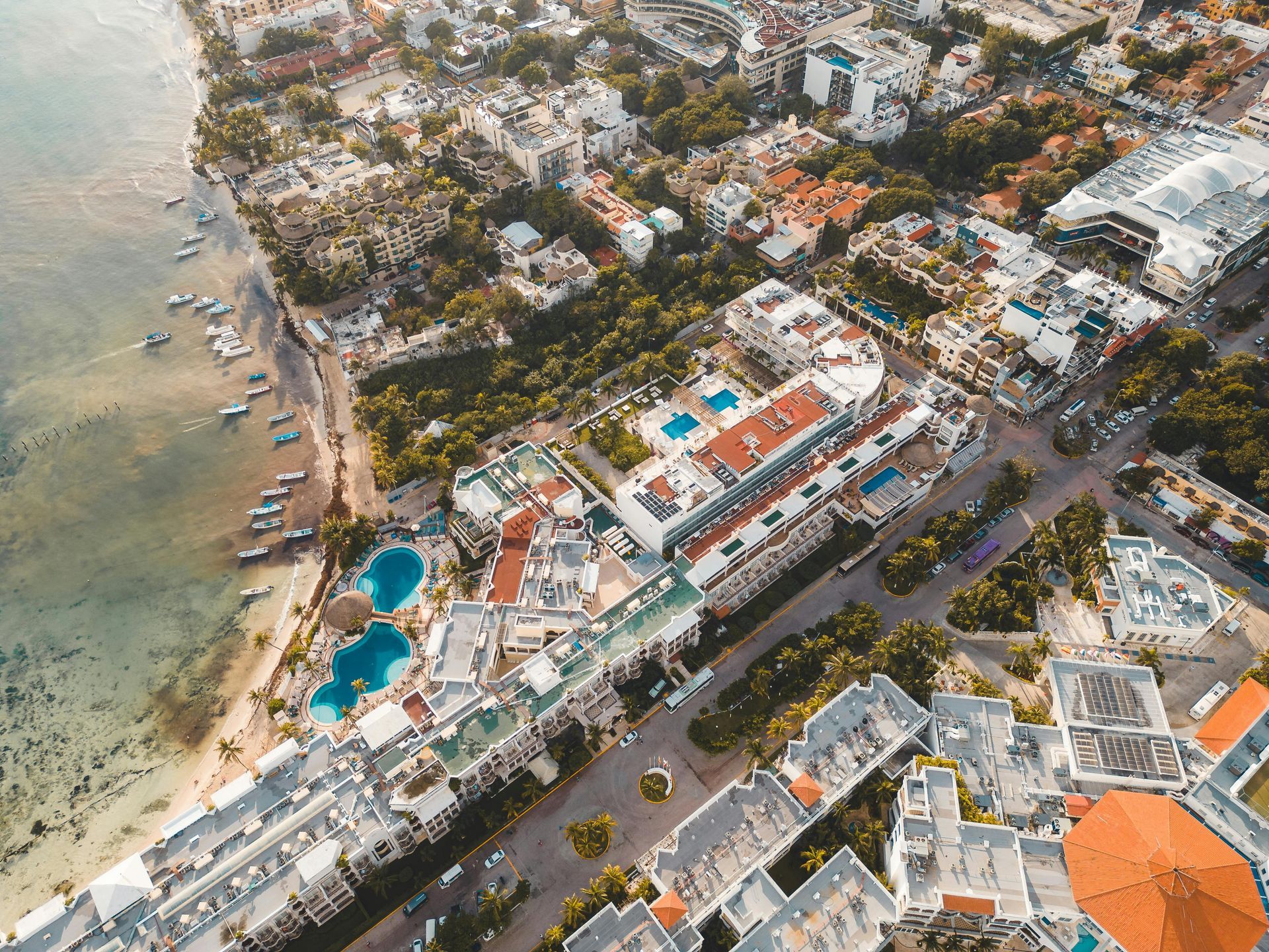 Aerial view of a coastal resort town with white buildings, blue pools, and a sandy beach along the turquoise water.