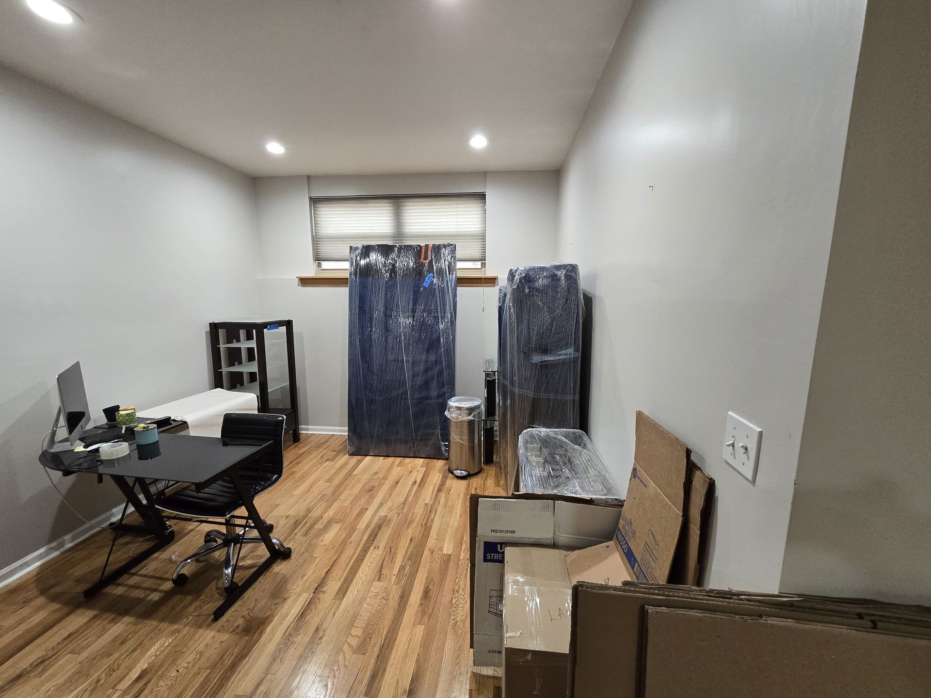 A room with wood floors, a desk and chair on the left, and several large items wrapped in blue plastic near the window.