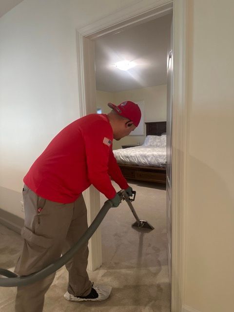 A man in a red shirt is using a vacuum cleaner in a bedroom.
