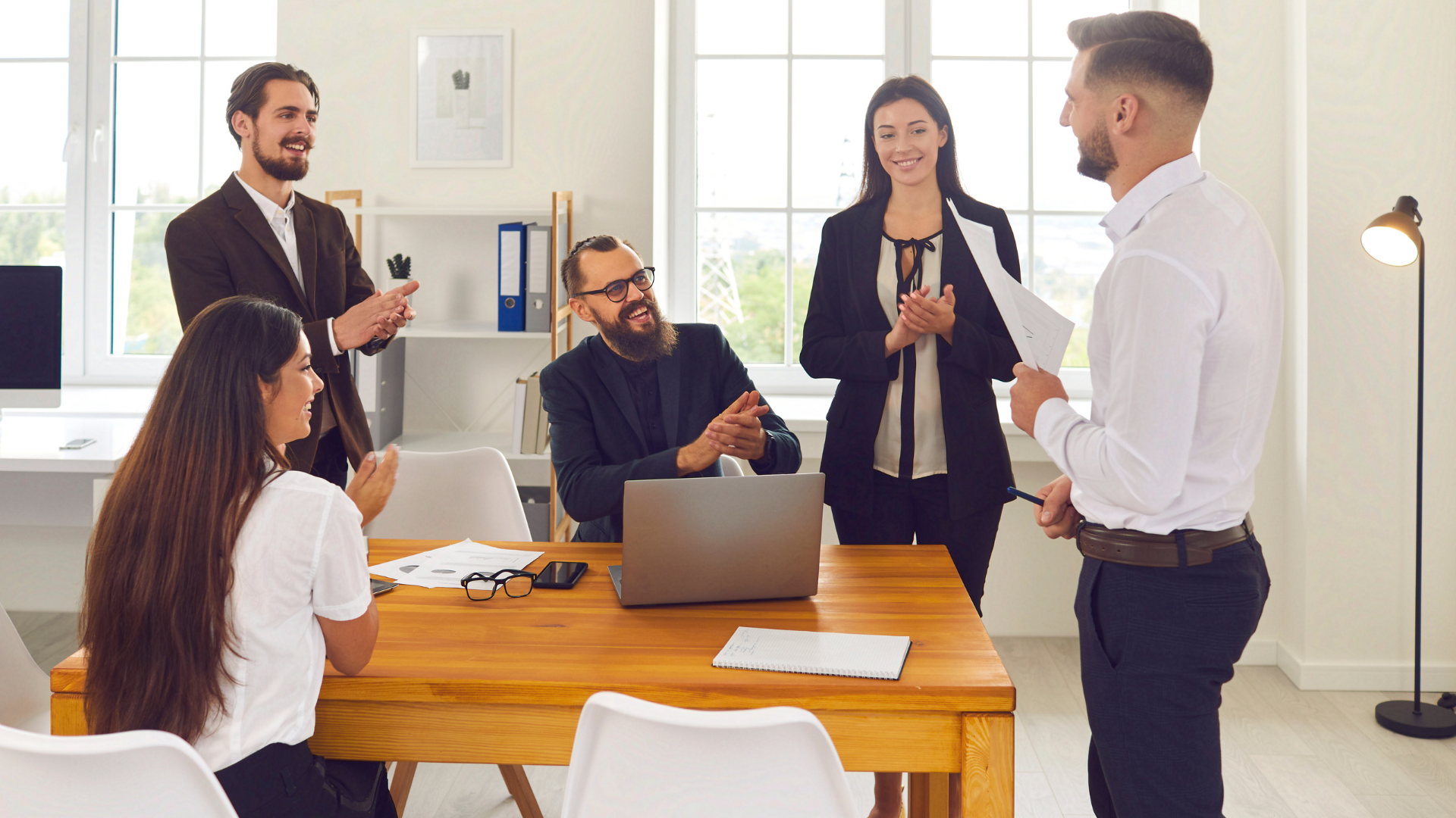 Office meeting: Team applauds a colleague presenting documents at a wooden table. Windows provide natural light.