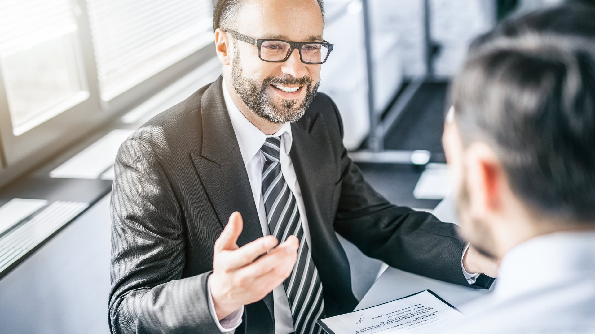 Man in suit smiles, gestures while speaking to another person in an office setting.