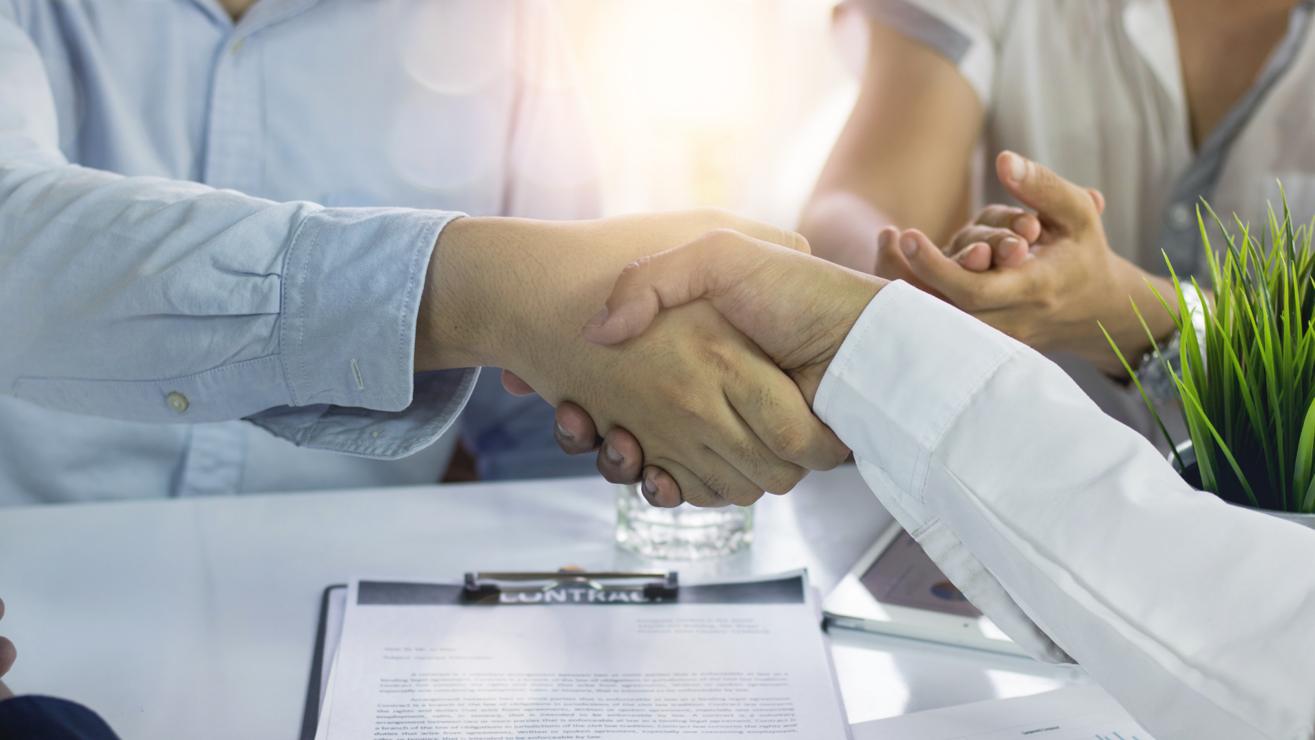 Two people shaking hands over a table with a clipboard and a plant.