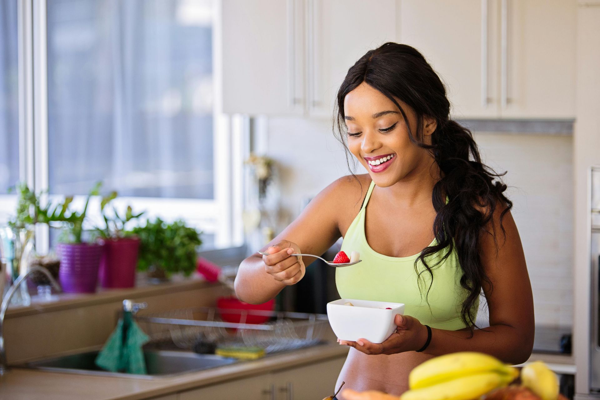 A person in a light green workout top eats a spoonful of berries while holding a small white bowl in a bright kitchen.