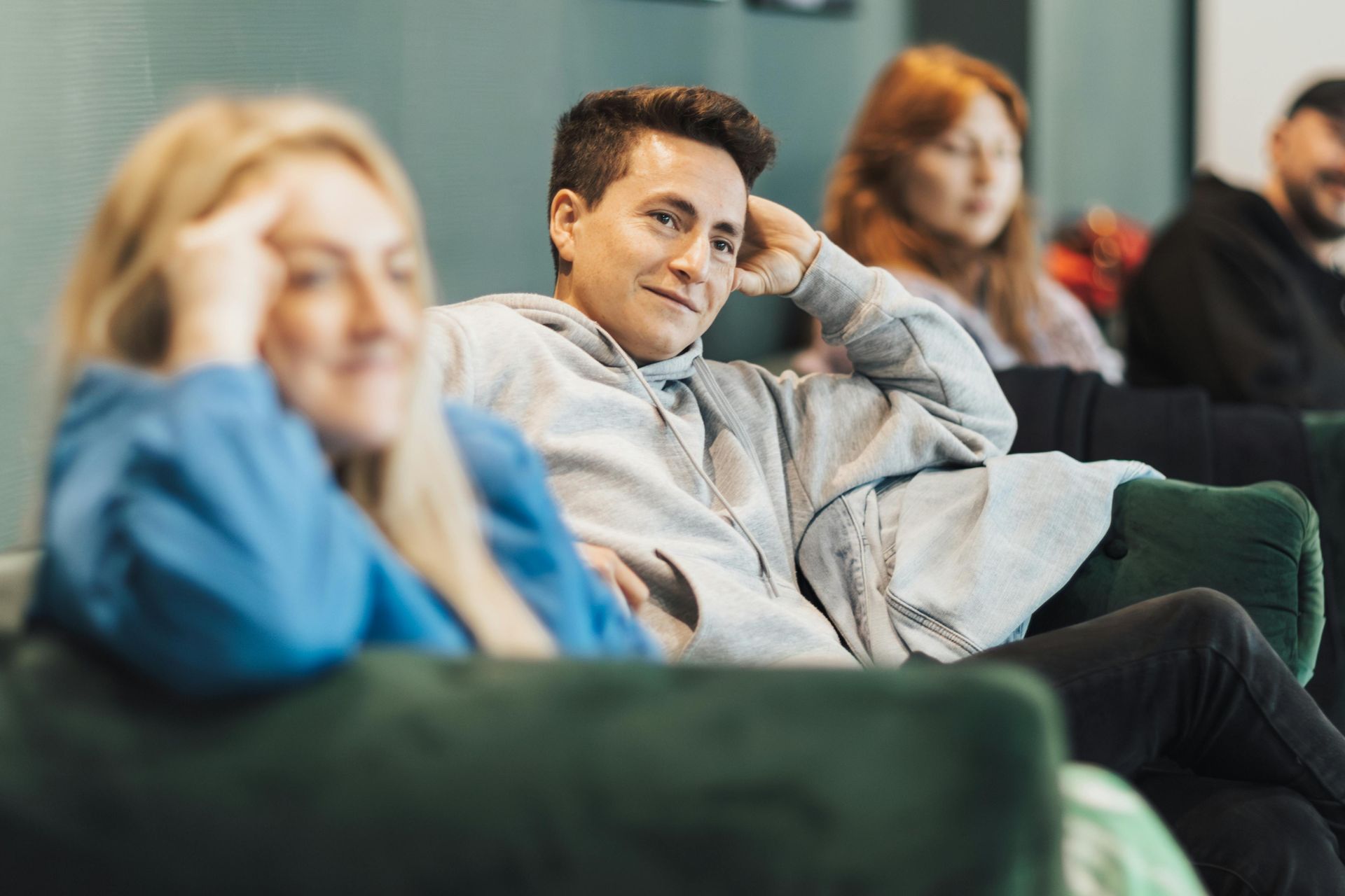 Three people sit on a green couch, with a person in a gray hoodie in the center looking toward the camera.