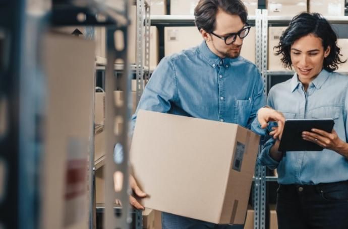 A man is holding a box and a woman is looking at a tablet in a warehouse.