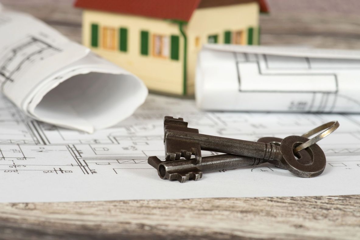 Due vecchie chiavi su piantine, progetti arrotolati e un modellino di casa su un tavolo, a simboleggiare la realizzazione di un immobile finito.
