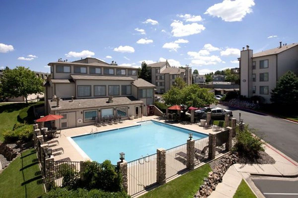 Swimming pool in front of a tan building under a blue sky with some clouds. Trees and a road.