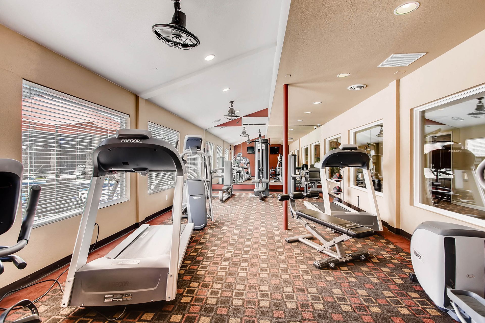 Gym with various exercise machines, windows with blinds, and mirrored wall.