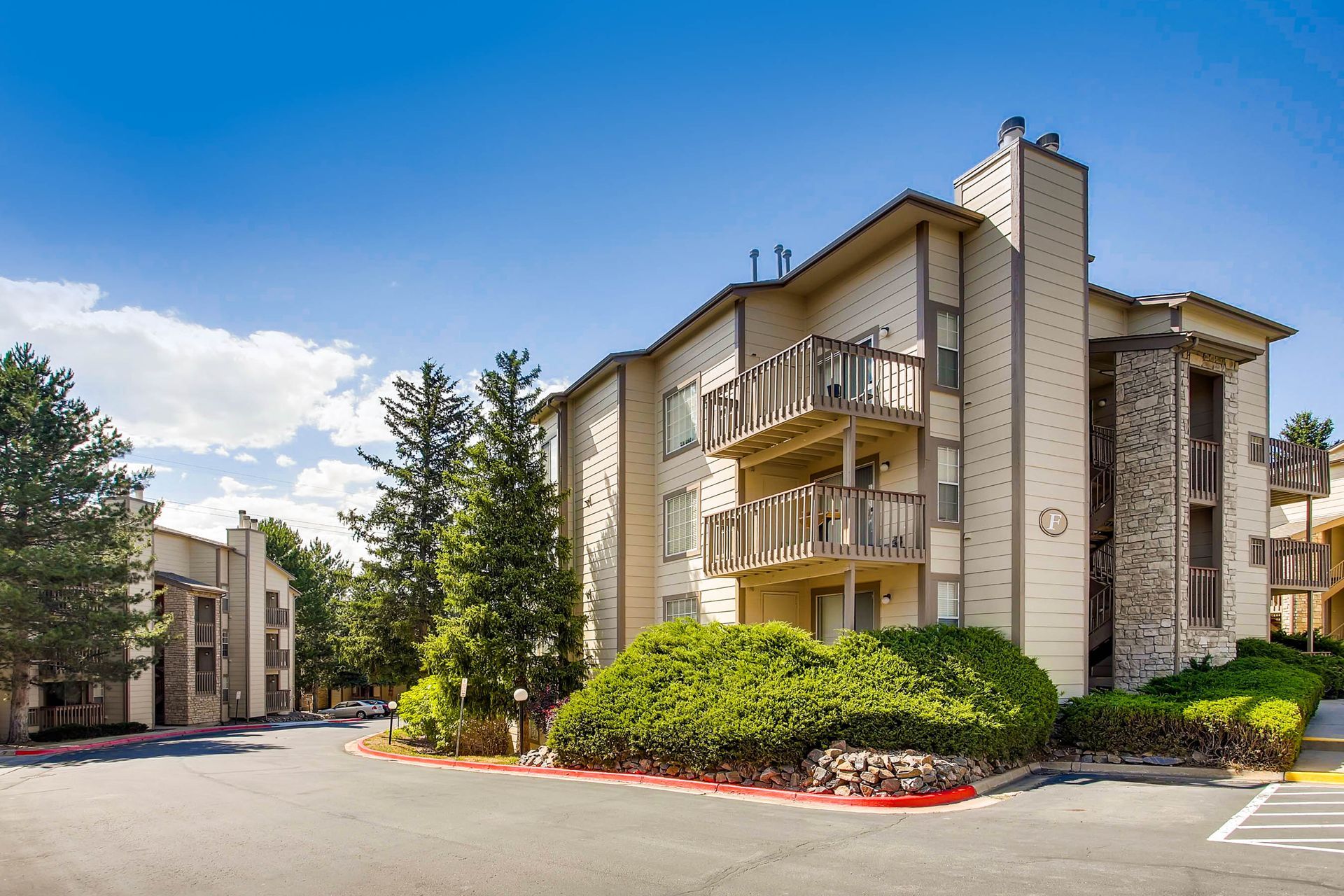 Apartment complex with beige siding, balconies, stone accents, and trees, against a blue sky.