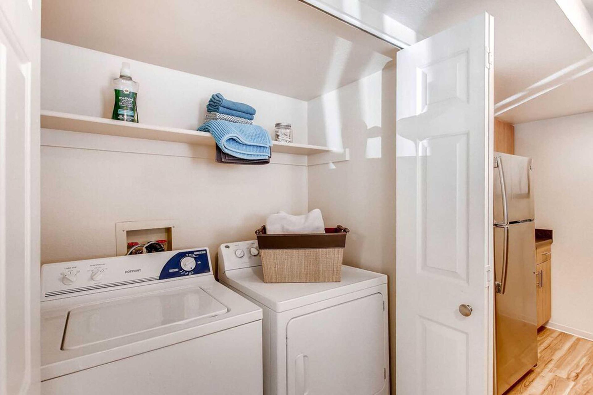 Laundry room with washer, dryer, shelves holding supplies, and an open door leading to a kitchen at Chestnut Ridge Apartment Homes, offers apartments near Denver Tech Center.