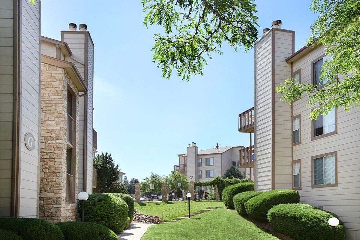Apartment buildings with beige siding and stone accents line a grassy walkway under a blue sky.