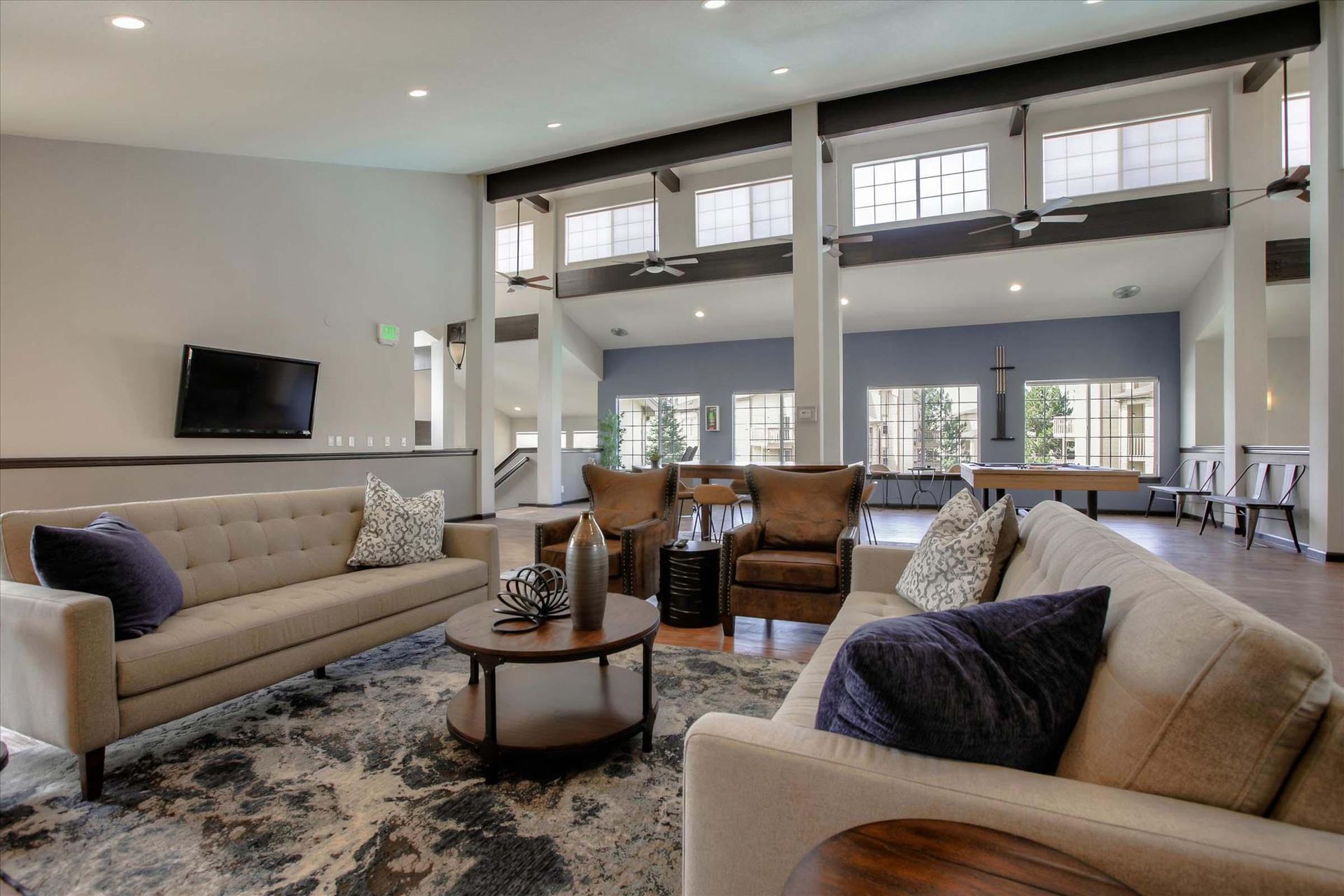 Lounge area with tan couches, round coffee table, area rug, and wall-mounted TV at Chestnut Ridge Apartment Homes, offers apartments for rent in Denver, CO. Large windows, open to other rooms.