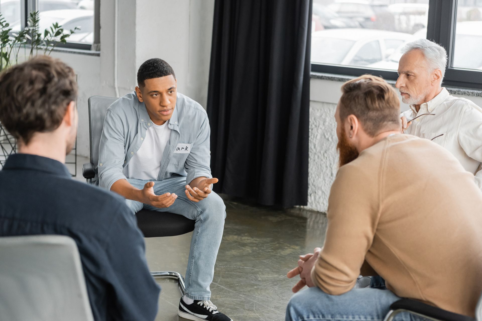 Men in a circle, one speaking. White and black men in casual clothes; natural light, indoor setting.