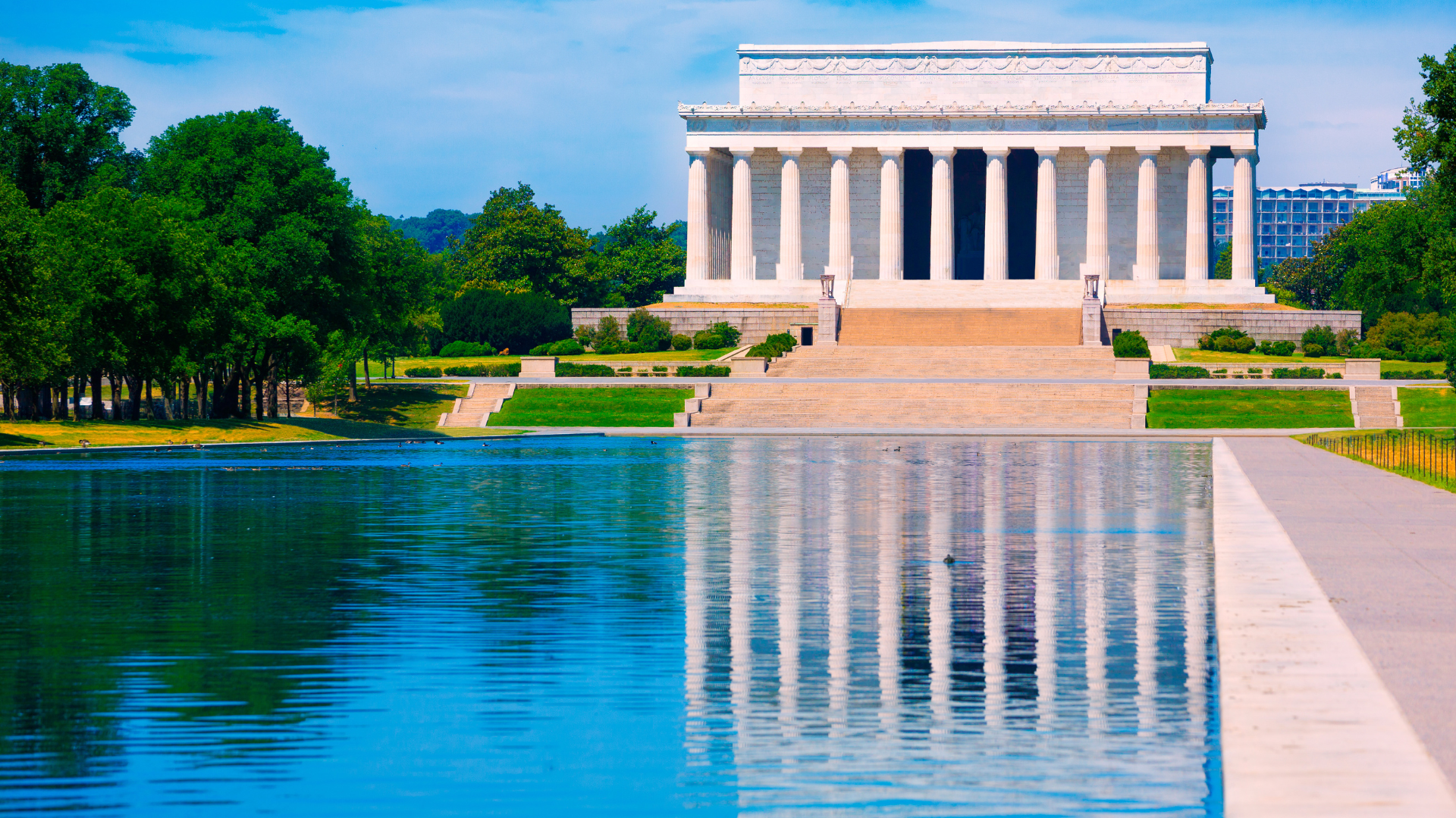 Lincoln Memorial reflecting in a long pool of water. White building with columns against a blue sky.