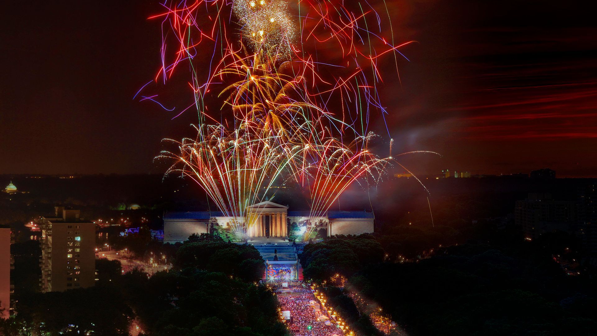 Fireworks erupt over a monument at night, illuminating a crowd below with red, gold, and blue.