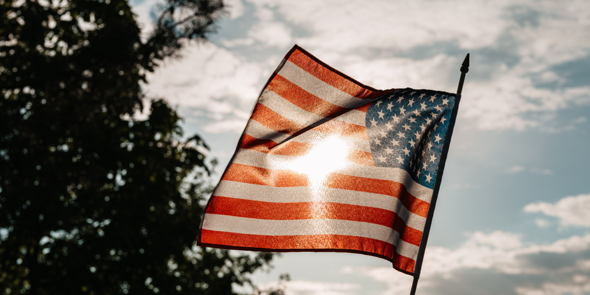 American flag waving against a bright sky, sunlight shining through.