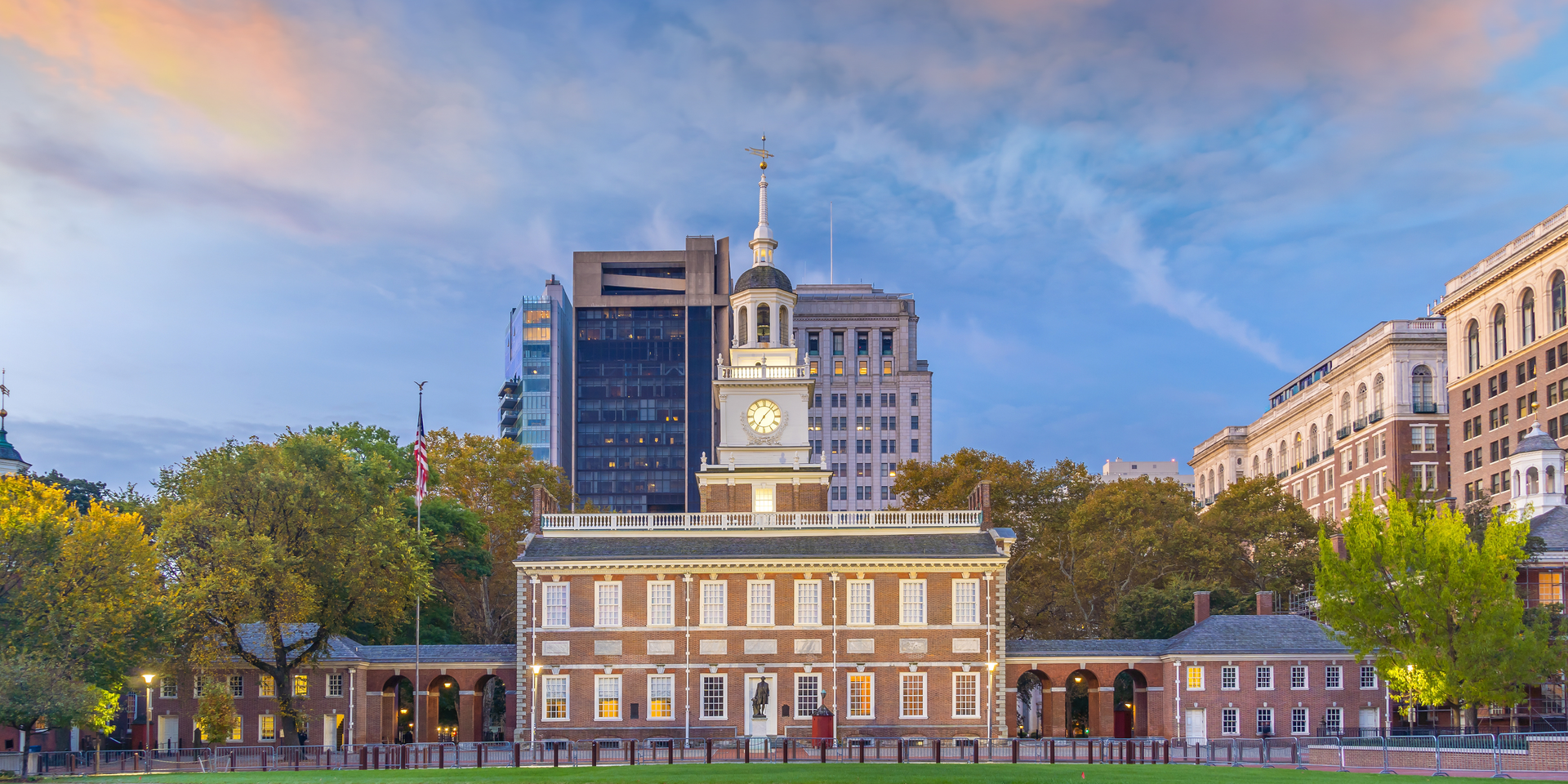 Independence Hall, Philadelphia, seen at dusk. Brown brick building with clock tower, surrounded by trees and other buildings.