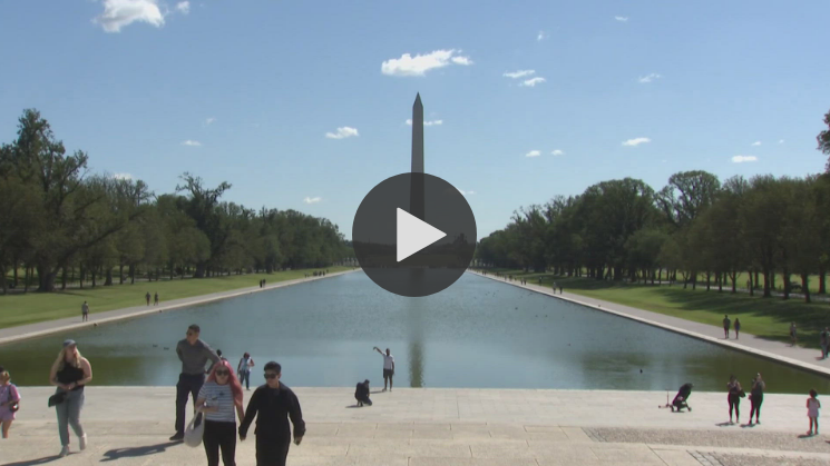 The Washington Monument, reflected in a long pool, with people on the plaza on a sunny day.