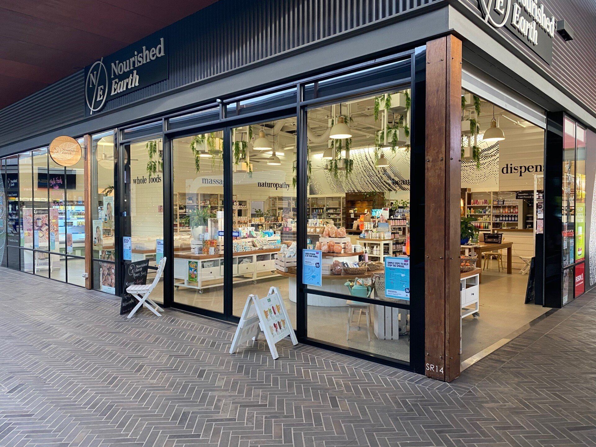 Corner storefront with large glass windows, black awning, and outdoor display signs on a paved sidewalk.