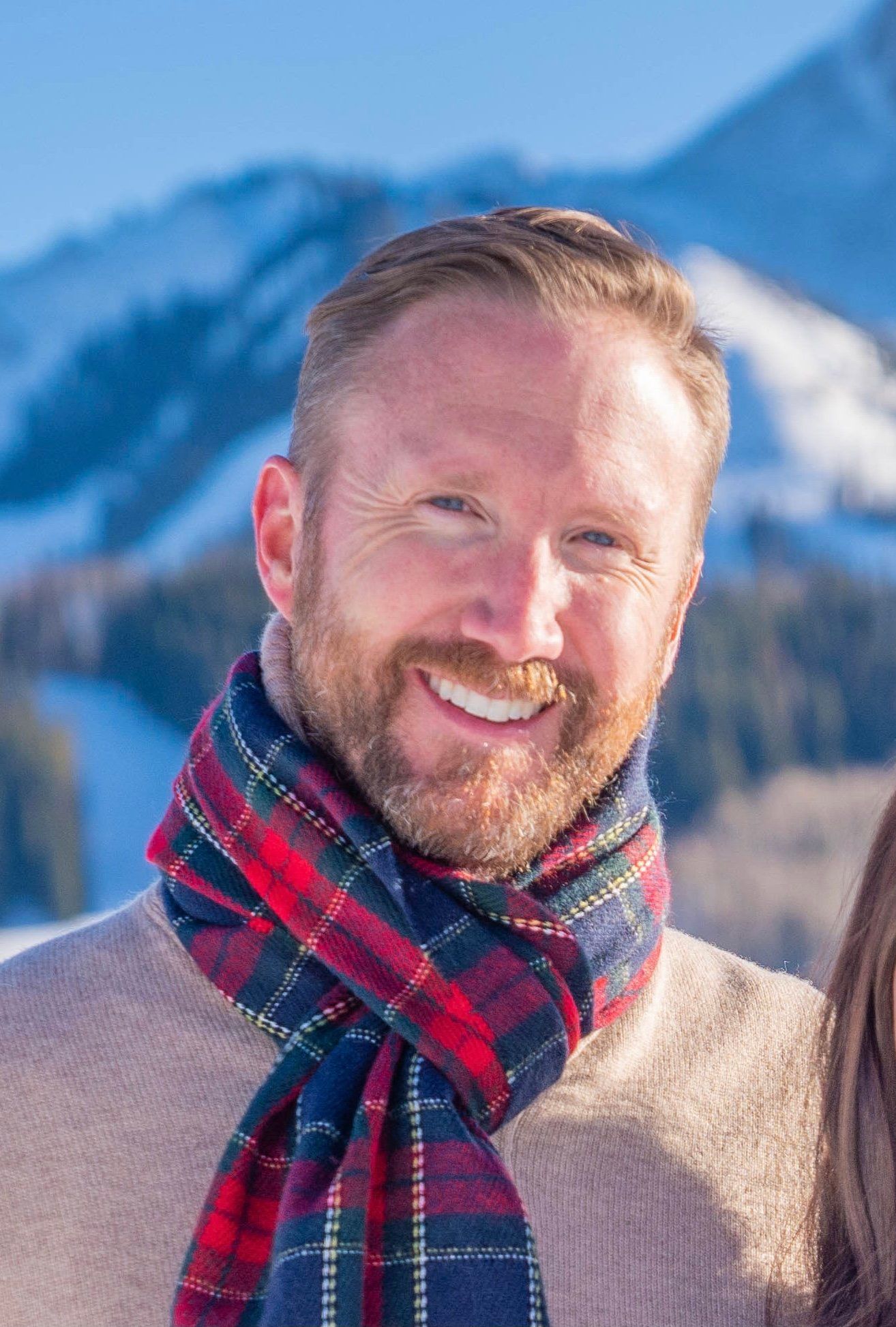 Man with red beard, wearing a plaid scarf, smiling in front of a snow-covered mountain.