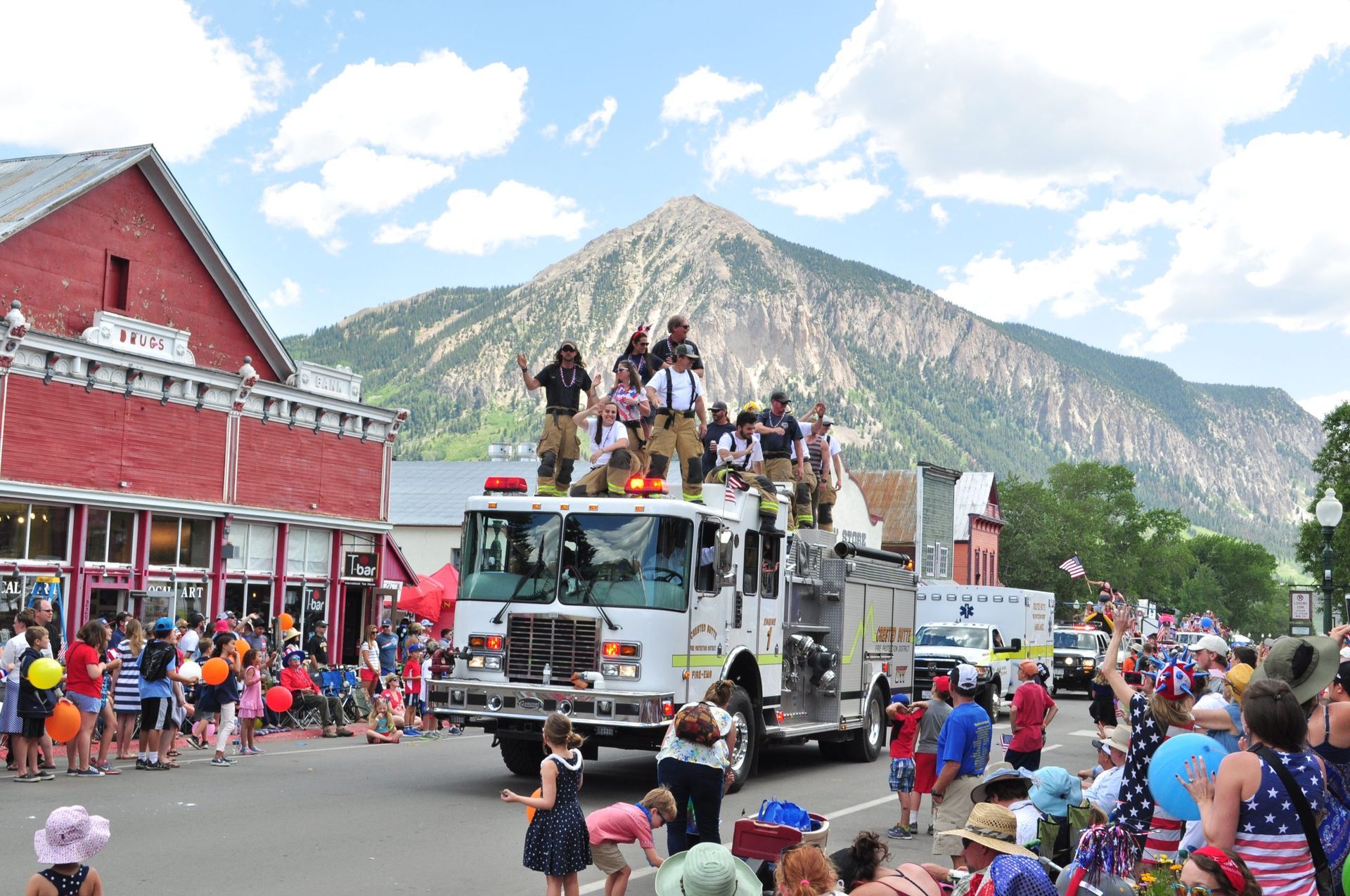 Firefighters on truck in parade, crowds on street, red buildings, mountain backdrop.