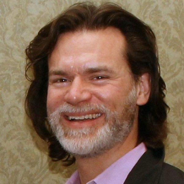 Smiling man with beard, long brown hair, wearing pink shirt and dark blazer.