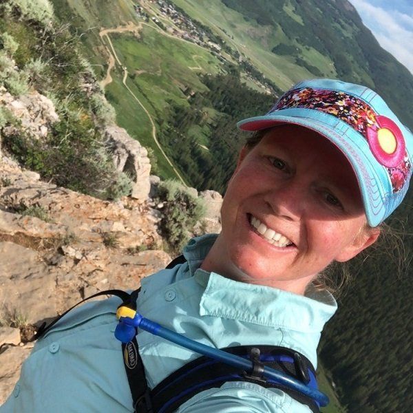 Woman smiles, wearing a cap and shirt, hiking on a mountain trail, with a scenic background.