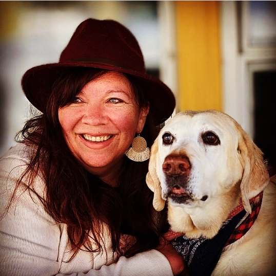 Woman in hat smiling next to a yellow lab with a red bandana.
