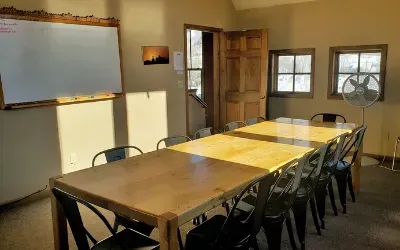Meeting room with a long wooden table, black chairs, windows, and whiteboard; sunny interior.