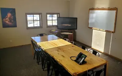 Conference room with a long wooden table, chairs, TV, windows, and whiteboard.