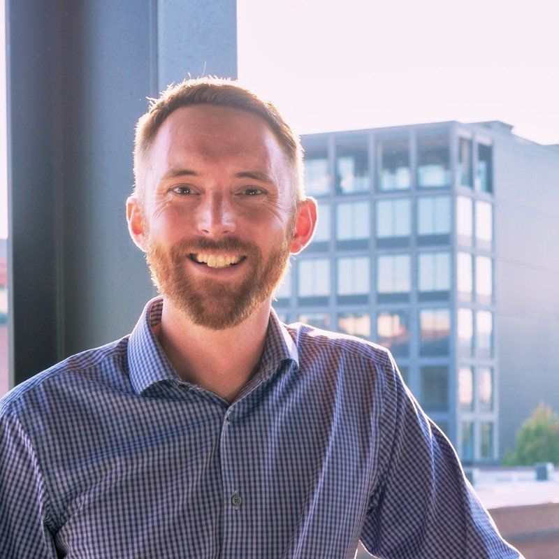 Man smiling, wearing blue plaid shirt, standing on balcony with city buildings in the background.