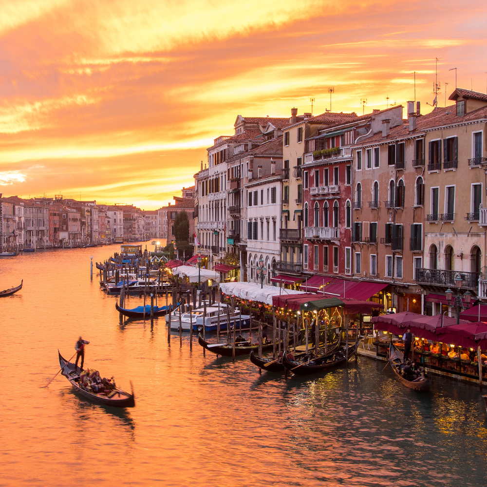 A gondolier navigates a boat on the Venice Grand Canal at sunset, lined by historic buildings and waterside restaurants.