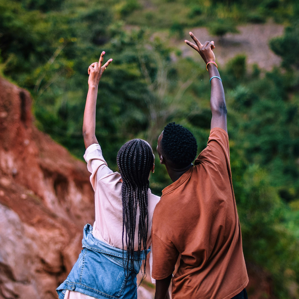 Two people seen from behind stand outdoors, arms raised in a V-sign against a backdrop of green hills and red earth.