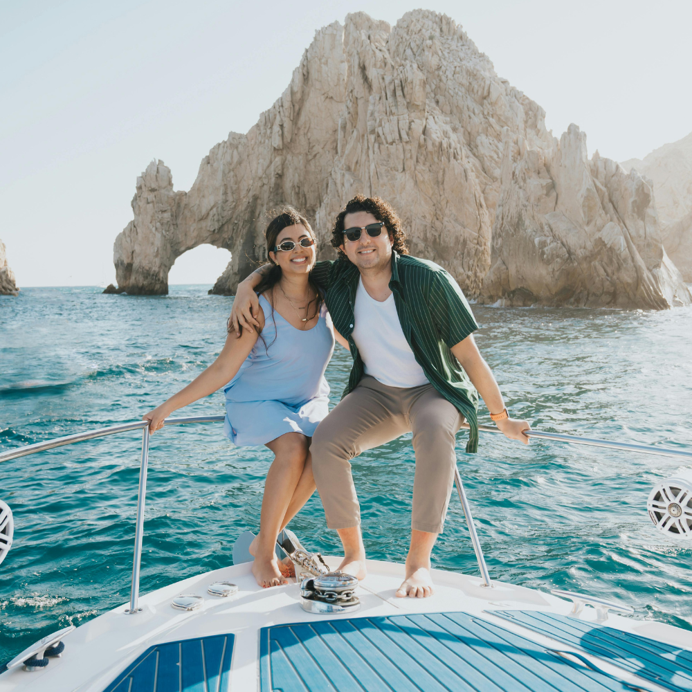 A couple sits on the bow of a boat in front of the iconic Arch of Cabo San Lucas, smiling toward the camera.