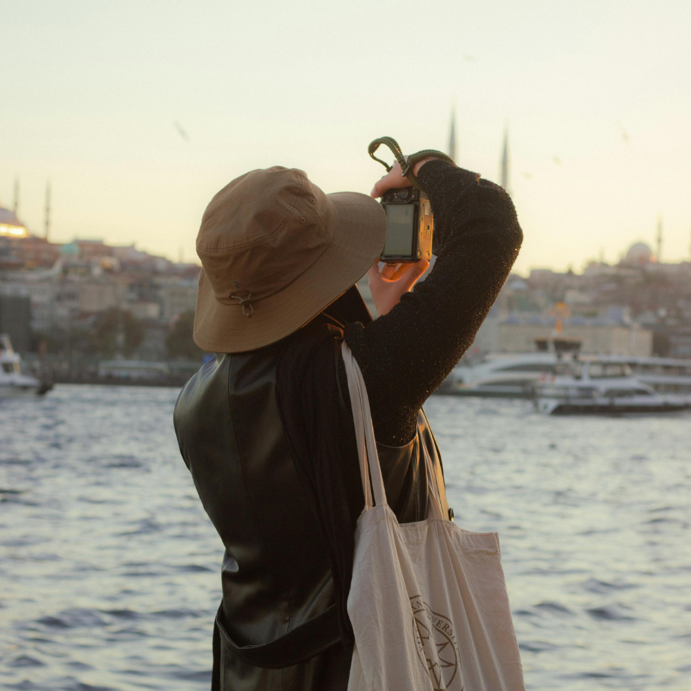A person in a bucket hat and dark coat takes a photo of a coastal cityscape with a camera, carrying a white tote bag.