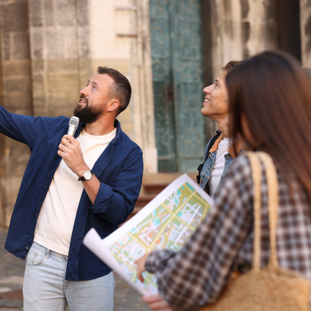 A tour guide with a microphone gestures toward a building while pointing out sights to two tourists holding a map.
