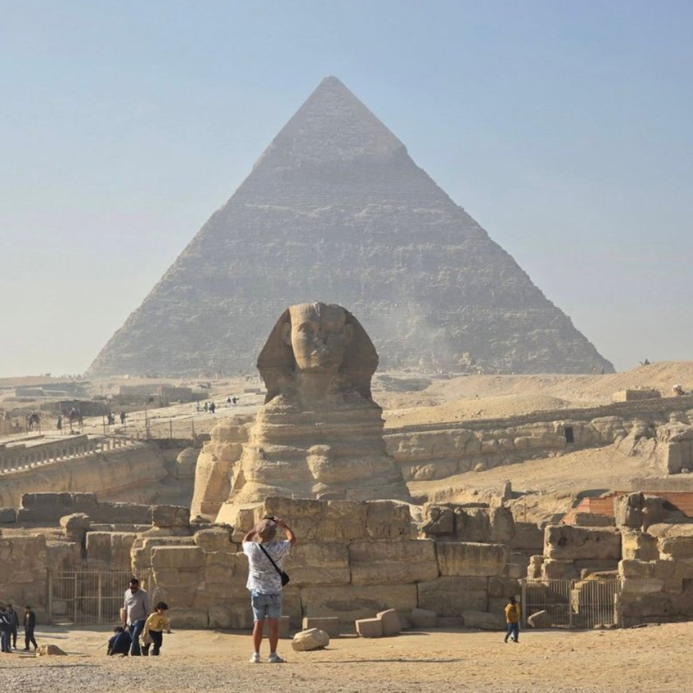 The Sphinx and the Great Pyramid of Giza stand in a desert landscape under a clear sky, with people visiting the site.