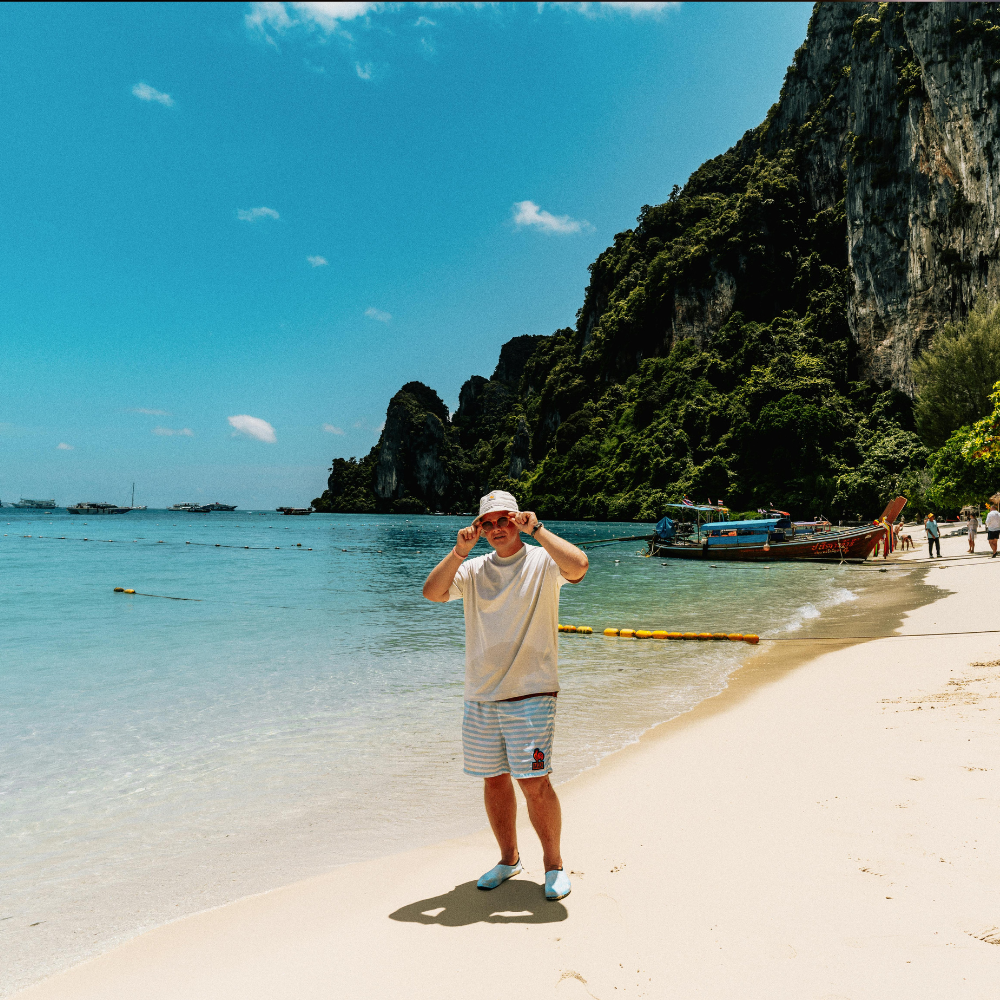 A person wearing a hat and light clothing stands on a sunny, sandy beach with cliffs and a boat in the background.