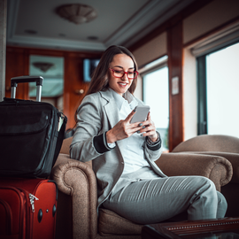 A traveler in a grey suit and red glasses sits in a lounge chair using a phone, with luggage nearby.