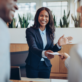 A smiling professional in a dark blazer hands a document to a client across a wooden office desk.