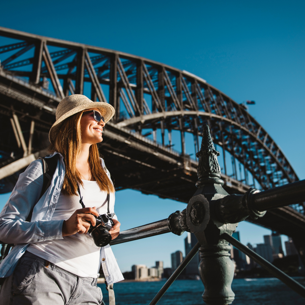 A smiling person in a sun hat holds a camera in front of the Sydney Harbour Bridge against a clear blue sky.