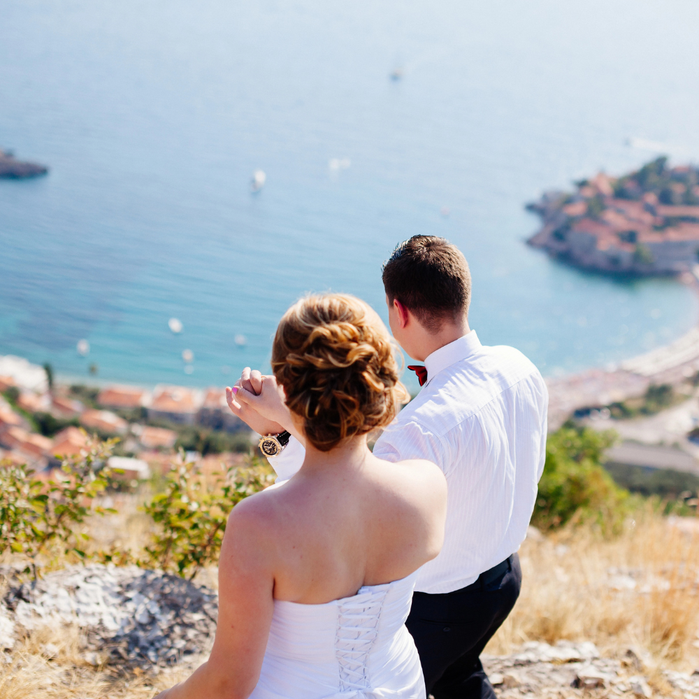 A bride and groom in wedding attire hold hands while overlooking a coastal town and sea from a grassy hillside.