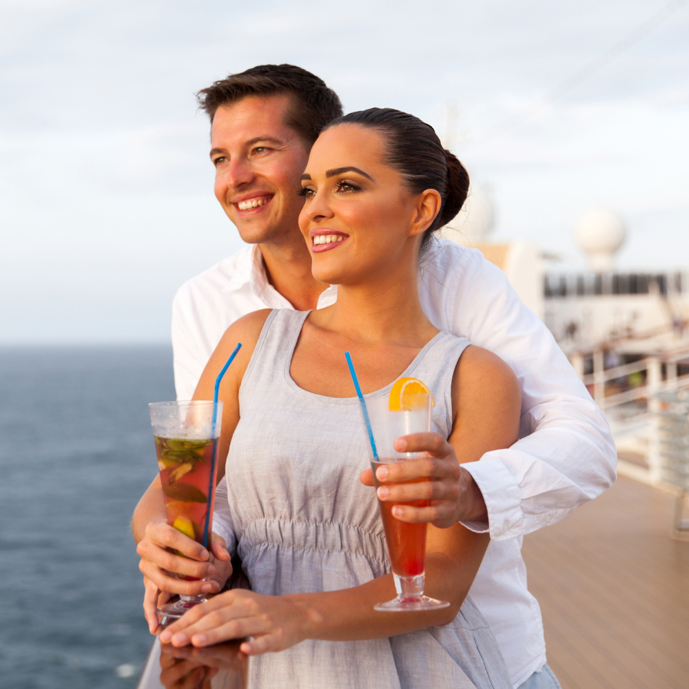 A couple stands on a cruise ship deck holding cocktails, smiling and looking out at the ocean.