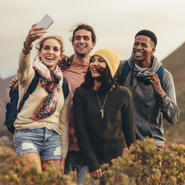 Four friends smiling and taking a selfie outdoors in a hilly, natural setting, wearing casual hiking attire.