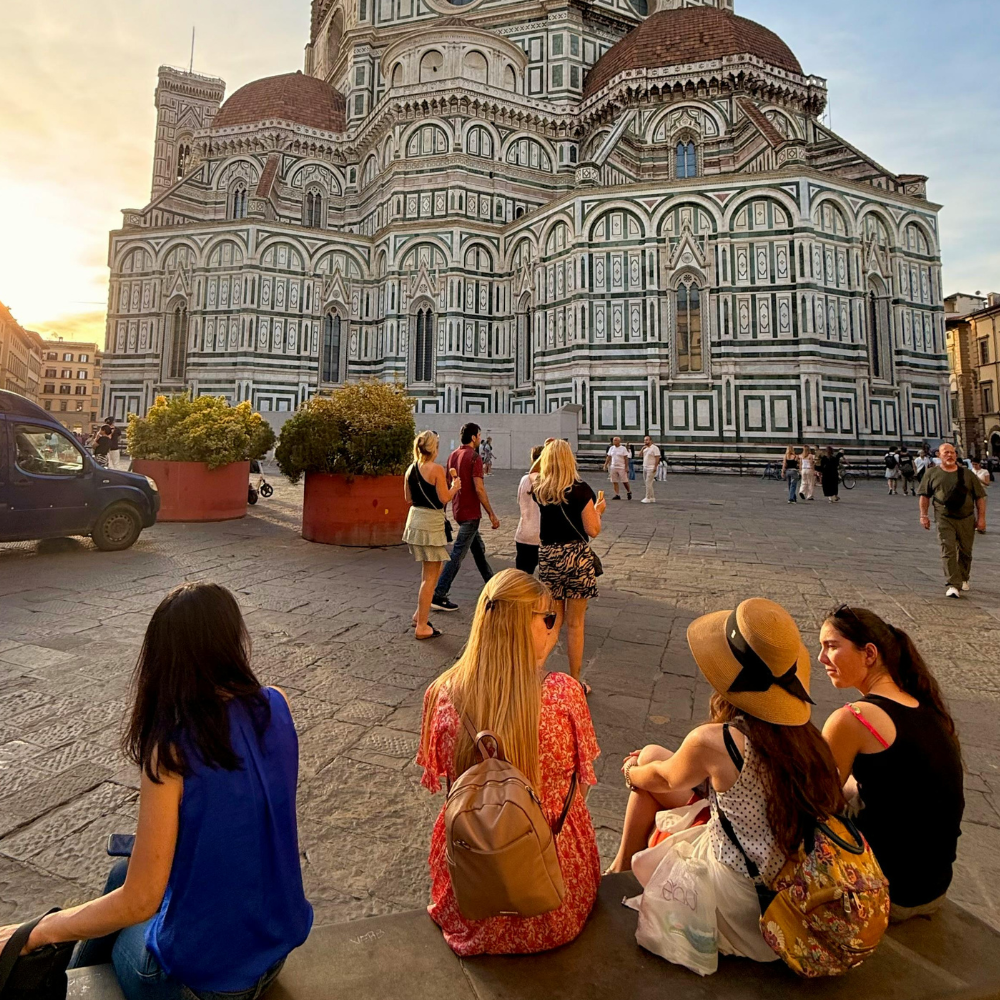 Four people sit on stone steps looking toward the Cathedral of Santa Maria del Fiore in Florence at sunset.