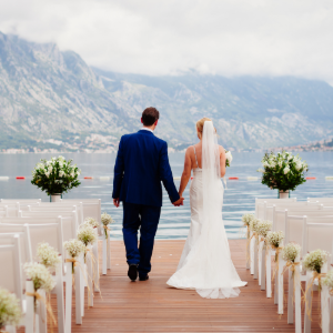 A couple holding hands and walking down an aisle lined with white chairs and flowers toward a lake and mountains.